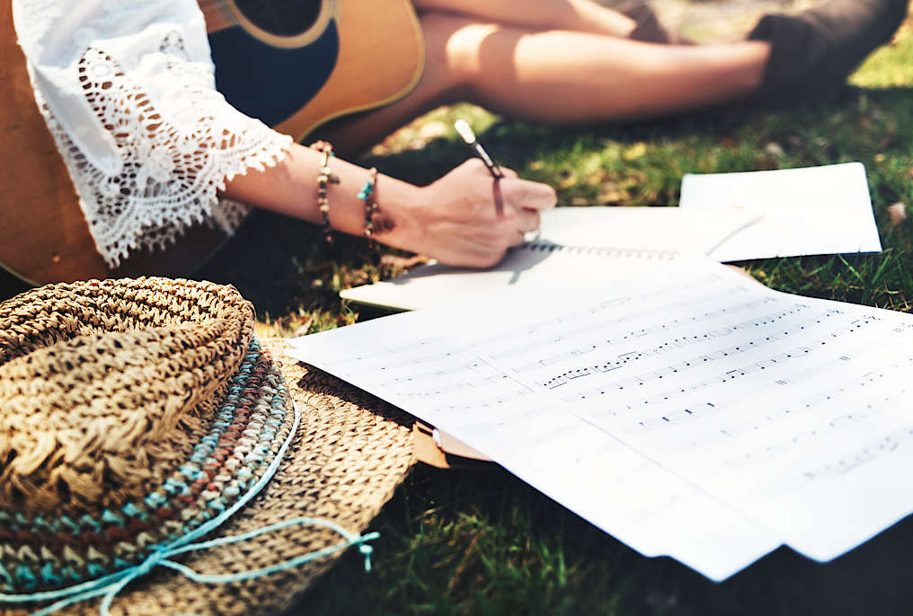 Someone sitting on grass on a sunny day, holding an acoustic guitar and writing sheet music
